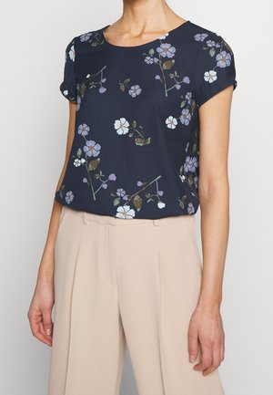 Vrouw draagt een donkerblauwe kortemouwtop met paarse en witte bloemenprint, ingestopt in beige broek met hoge taille.