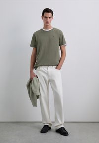Jonge man in een olijfgroen T-shirt boven een wit onderhemd, witte broek, zwarte schoenen, met een bijpassende olijfgroene gebreide trui in de hand, staande binnen.