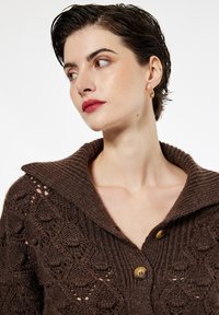 Brązowy sweter z dzianiny z szerokim kołnierzem, teksturowanym wzorem i dwoma drewnianymi guzikami przy dekolcie.