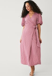 Rosa Wickelkleid aus strukturiertem Stoff, mit kurzen Puffärmeln, einem Bindegürtel und einem mittellangen Saum, der ein dezentes, zerknittertes Muster aufweist.