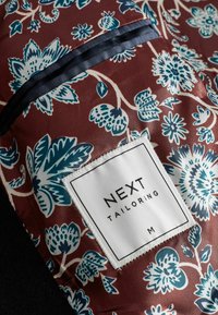 Burgundinės, mėlynos ir baltos spalvos gėlių rašto audinys su prisiūta balta etikete, kurioje parašyta "NEXT TAILORING M."