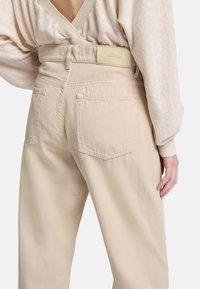 Beige hoog-getailleerde broek met een rechte snit, voorzien van twee achterzakken en een minimalistisch taillelabel. Zachte stoftextuur.