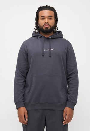 Man with braided hair and beard wearing a dark gray hoodie with front pocket and drawstrings, standing against a plain light background.