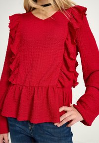 Röd texturerad blus med volangdetaljer, V-ringning och peplumslut. Långa ärmar med utsvängda muddar. Parat med blå jeans.