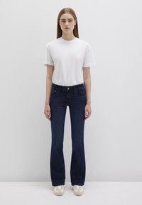Dark blue flared jeans, cotton blend, five pockets, mid-rise waist. Paired with a plain white crew-neck t-shirt and light-colored sneakers.