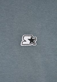 Tissu gris avec un petit logo brodé blanc et noir présentant une étoile et une lettre « S » stylisée au centre.