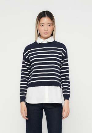 Maglione corto a righe navy con righe bianche, texture a maglia, scollo rotondo, abbinato a una camicia bianca con colletto sotto e jeans scuri.