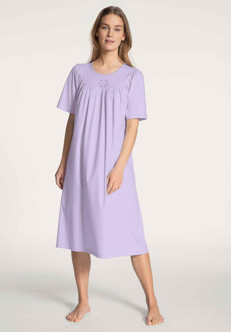 Camicetta in lavanda in morbido tessuto con maniche corte, scollatura rotonda e dettagli arricciati sul corpetto. La lunghezza arriva a metà polpaccio.