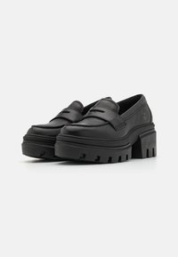 Musta nahkainen platform-loaferit leveällä hihnalla, tukeva pohja ja teksturoitu yksityiskohta. Pyöreä kärki ja huomaamaton logo kantapäässä.