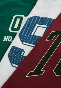 Drie t-shirts die elkaar overlappen: groen met witte tekst, wit met blauw gemusterde cijfers, en kastanjebruin met zwart omlijnde cijfers. Katoenen materiaal.