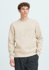 Beige sweatshirt met lange mouwen, ronde hals en een voorzak. Zachte textuur, eenvoudig ontwerp en een relaxte pasvorm.
