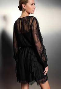 Robe noire en dentelle avec un ourlet à volants et de longues manches transparentes. Le dos présente un détail en forme de trou de serrure avec une fermeture à bouton. Tissu texturé.