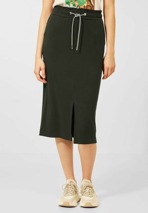 Czarna spódnica do kolan z przednim rozcięciem i ściągaczem w talii, noszona z beżowymi trampkami, na stojącej osobie.