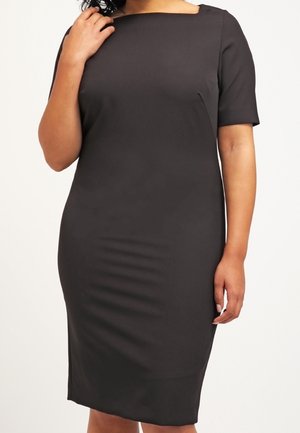 Kobieta w dopasowanej, czarnej sukience o długości do kolan z krótkimi rękawami i dekoltem typu łódka, stojąca z jedną ręką przy twarzy.