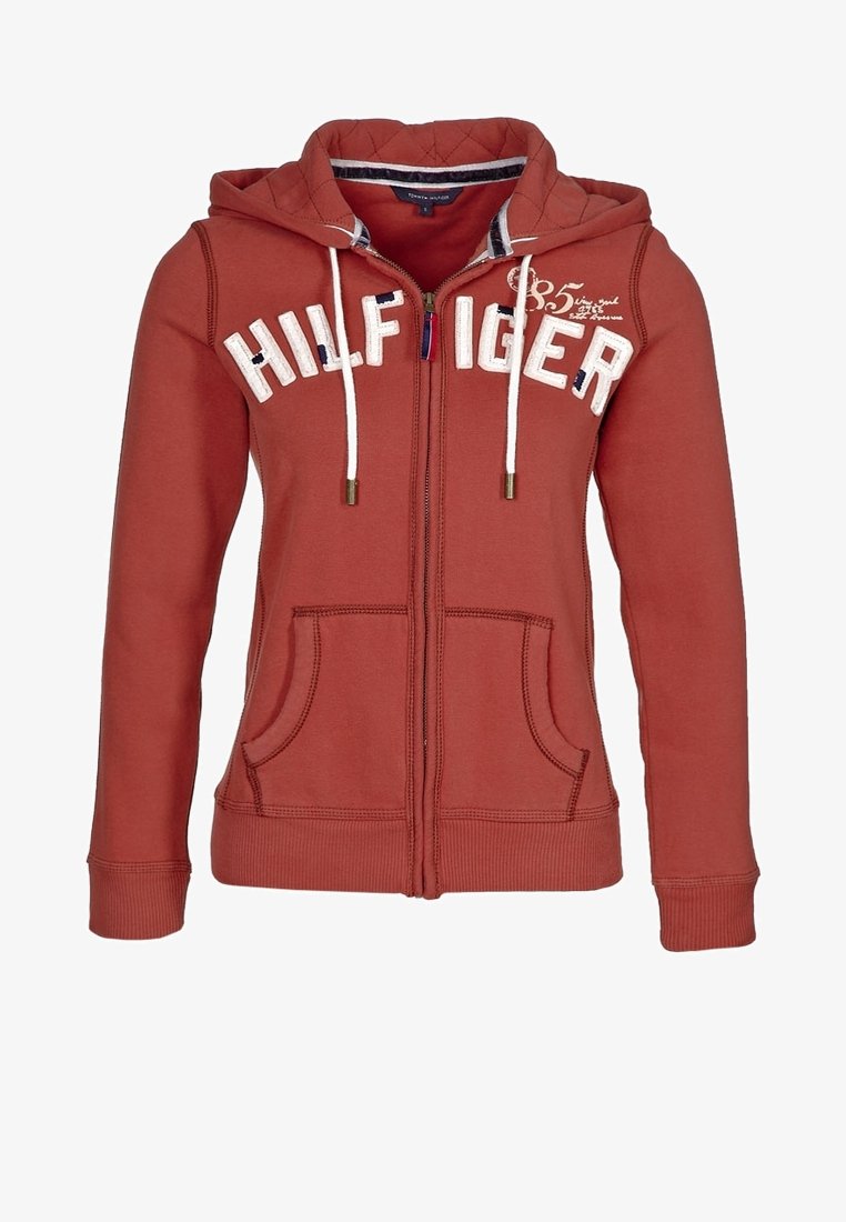 Sweat à capuche zippé couleur rouille avec cordons blancs, poches avant et texte "HILFIGER" sur la poitrine.