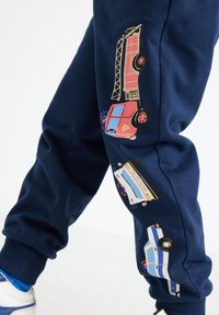 Marineblå joggingbukser med farverige, tegneserieagtige prints af nødkøretøjer på siden, med elastiske manschetter og en blød tekstur.