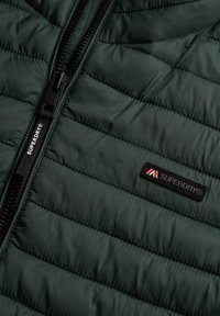 Ciemnozielona pikowana kurtka z czarnym zamkiem błyskawicznym oraz czatnym prostokątnym logo z napisem "SUPERDRY®" w kolorze białym i czerwonym.