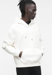 Sudadera con capucha color crema con bolsillo frontal, logo en bordado claro y puños oscuros a contraste. Bolsillo tipo canguro en la parte delantera.