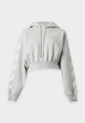 Femme portant un sweat à capuche gris court et un pantalon de survêtement noir avec des baskets blanches, debout devant un fond uni clair.