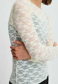 Haut en dentelle avec des motifs floraux crème et bleu clair, manches longues en voile et coupe ajustée. Tissu texturé et léger.