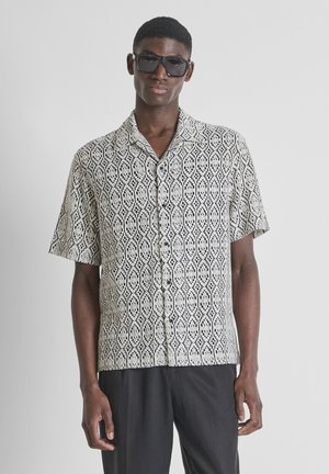 Joven con gafas de sol oscuras y una camisa de manga corta estampada en blanco y negro con pantalones negros sobre un fondo liso.