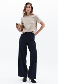 Beige kortärmad topp och marinblå vida byxor. Modellen håller en brun väska och bär svarta klackskor med guldaccenter. Slät textur.