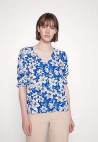 Blusa floral azul com mangas curtas em balão, decote em V e detalhe de botões. Apresenta padrões de flores brancas e pretas sobre um fundo azul vívido.