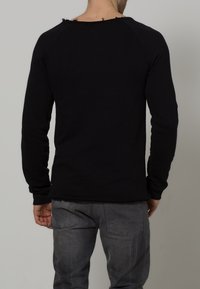 Chemise noire à manches longues avec un col brut, au design ajusté et en tissu texturé, portée par une personne en jean gris.