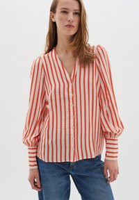 Blouse rayée rouge et blanche avec un décolleté en V, fermeture à boutons et manches bouffantes. Tissu léger et texturé. Portée avec un jean en denim bleu.