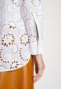 Chemisier blanc brodé avec des motifs floraux découpés, manches longues et poignets à boutons, associé à une jupe en cuir marron.