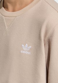 Bež pulover z vzorcem iz kvačkane vaflje, z rebrastim izrezom okoli vratu in izvezenim belim logotipom Adidas na levi prsni strani. Teksturiran material zagotavlja edinstven videz.