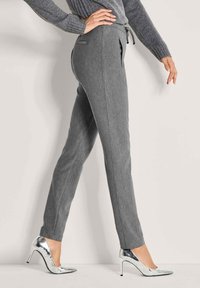 Vrouw in strakke grijze broek met zijzakken en zilveren hoge hakken, poserend tegen een lichte achtergrond.