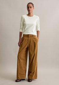 Frau steht mit den Händen in den Taschen, trägt ein weißes Langarmshirt, braune weite Cordhosen und dunkelbraune Schuhe.