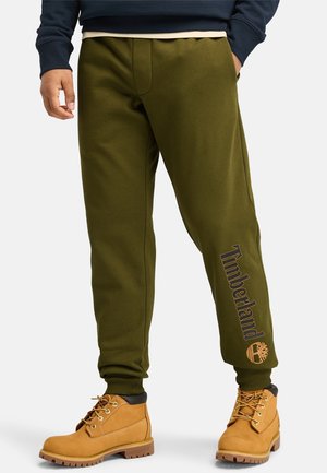 Mann trägt olivgrüne Timberland-Jogginghose mit Logo am Bein und sandfarbene Timberland-Stiefel, steht mit den Händen in den Taschen.