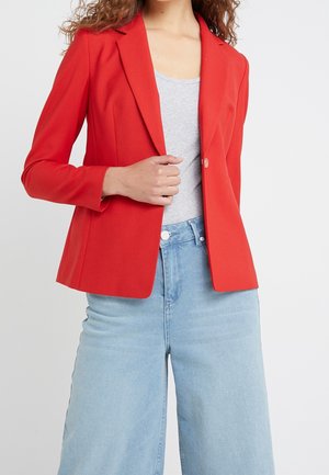 Frau trägt einen leuchtend roten Blazer über einem hellgrauen Oberteil und hoch taillierte, hellblaue, weit geschnittene Jeans, hält den Blazer-Revers.
