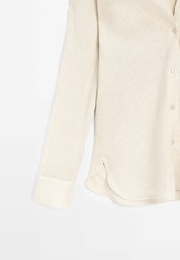 Chemise beige à manches longues avec boutons, ourlet arrondi et poignets boutonnés, vue de côté sur fond blanc.