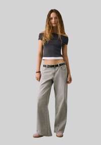 Top scurt negru cu umeri descoperiți, blugi largi gri, curea neagră cu ținte, încălțăminte roz, stând în fața unui fundal gri.