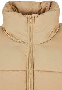 Beige pufferjakke med høy krage, med sentral glidelås og kviltet design. Materialet virker jevnt med en myk tekstur.