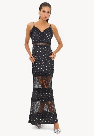 Femme portant une robe nuisette à pois noirs et blancs avec des panneaux en dentelle, des bretelles fines et des chaussures argentées à bouts pointus, debout devant un fond uni.