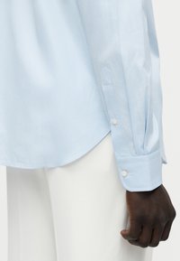 Άνδρας που φορά ανοιχτό μπλε πουκάμισο με μακριά μανίκια και ρίγες, με κουμπωμένα μανίκια και λευκό παντελόνι, ορατός από τη μέση μέχρι το χέρι.