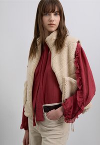 Chaleco sherpa crema con cuello alto sobre una blusa rojo oscuro con mangas con volantes, combinado con pantalones de pana beige claro.