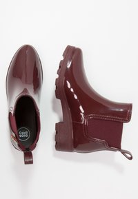 Bottes de cheville imperméables bordeaux avec une finition brillante, des panneaux latéraux élastiques et des semelles en caoutchouc texturé. Elles disposent d'une languette pour un enfilage facile.