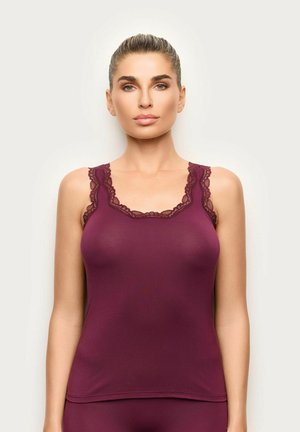 Mujer con el pelo recogido, vestida con una camiseta sin mangas burdeos con ribete de encaje, de pie frente a un fondo claro y liso.