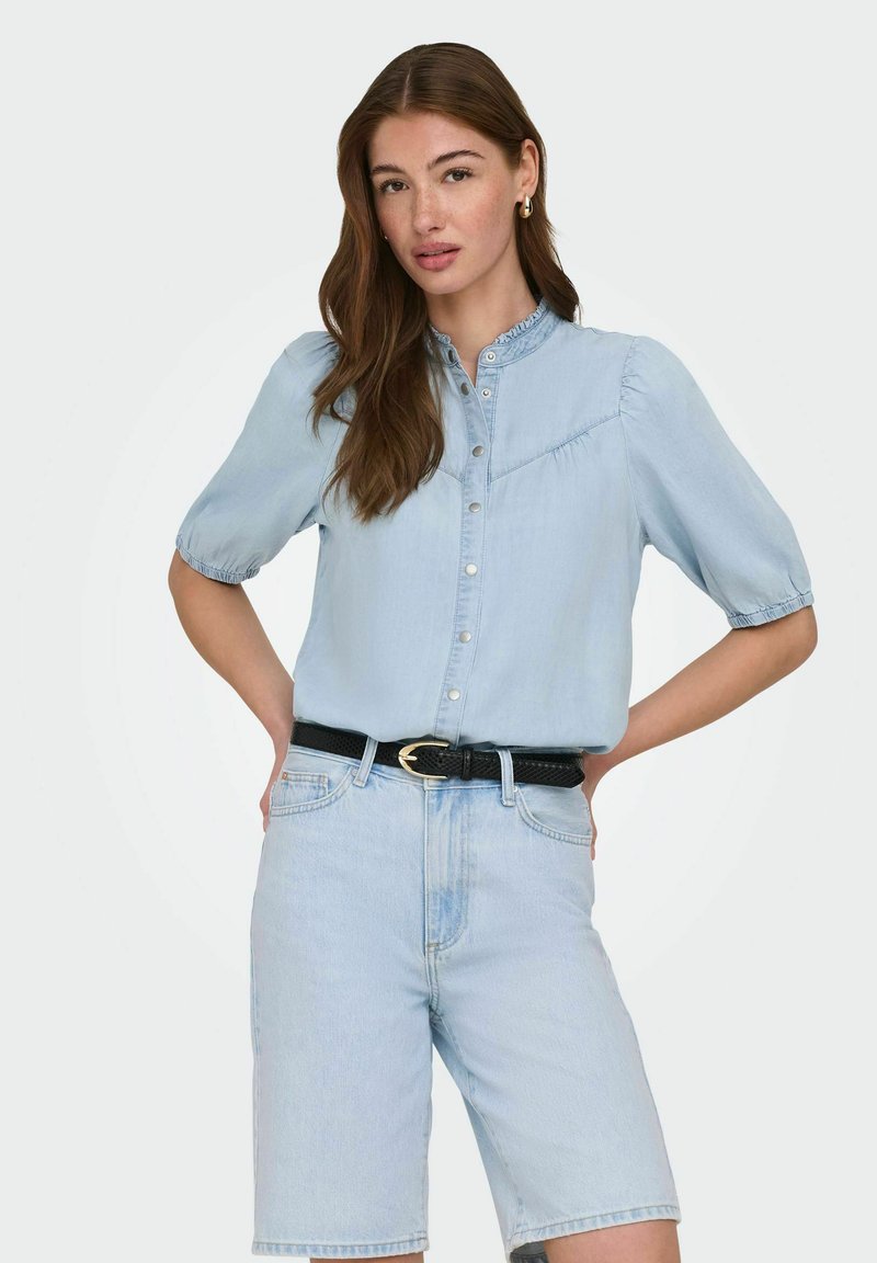 Kobieta o długich brązowych włosach nosi jasnoniebieską zapinaną na guziki bluzkę z bufkami oraz jasne dżinsowe szorty i czarny pasek, stoi z rękami na biodrach.