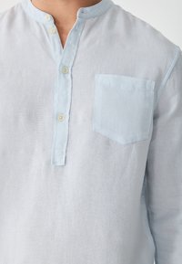 Light blue linen shirt with a mandarin collar, three buttons, and a front pocket. The fabric has a soft, textured appearance.