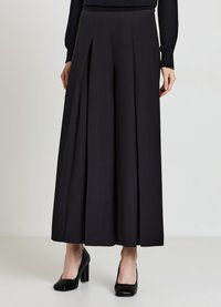 Donna che indossa pantaloni neri a vita alta e gamba larga con pieghe, abbinati a un top nero a maniche lunghe e scarpe nere con tacco block.