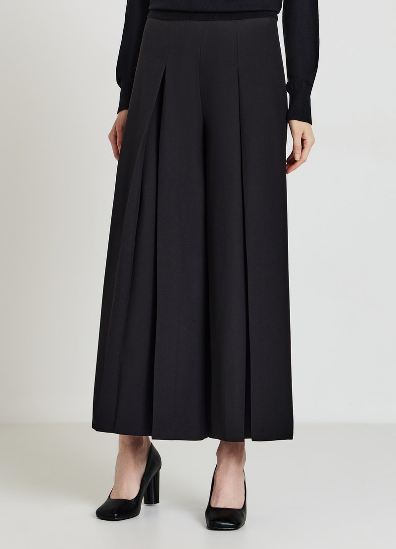 Donna che indossa pantaloni neri a vita alta e gamba larga con pieghe, abbinati a un top nero a maniche lunghe e scarpe nere con tacco block.