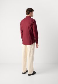 Bordeauxfarbene Hemdbluse mit Kragen, langen Ärmeln und glatter Textur, kombiniert mit cremefarbenen Cargo-Hosen und schwarzen Schuhen.