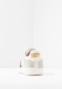 Zapatilla blanca y gris con acabado de ante, con el logo dorado "SUPERDRY" y rayas negras en el costado. Suela de goma y forro suave.