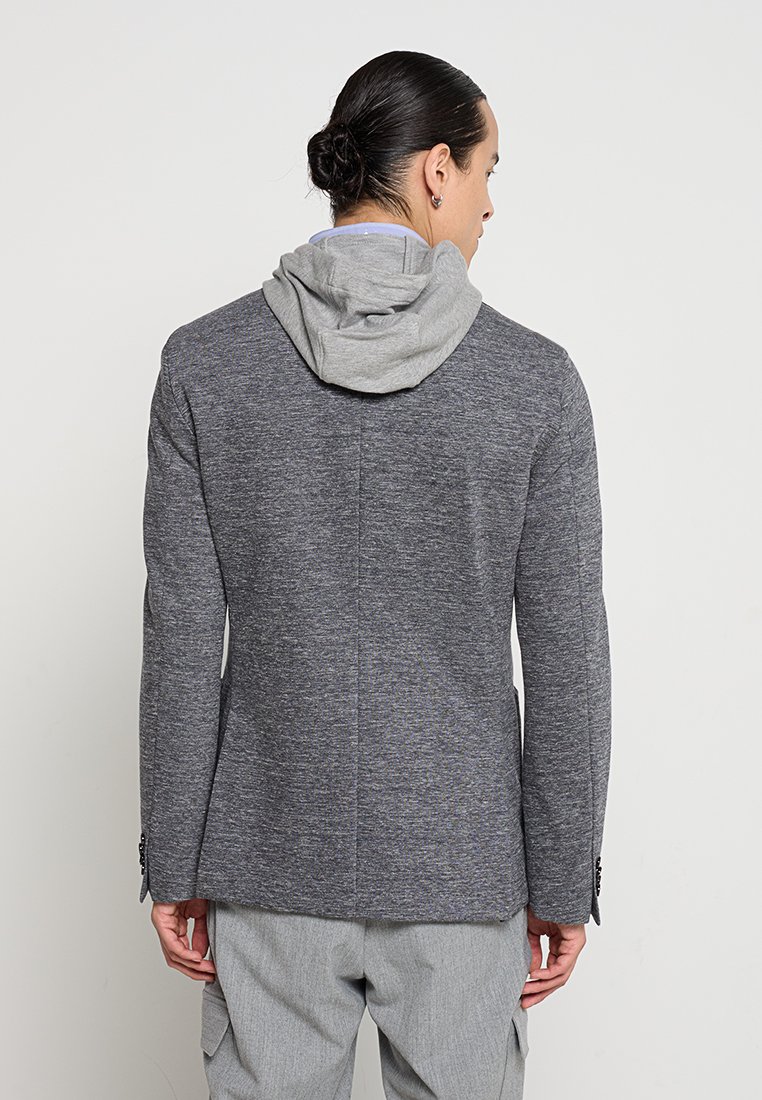 Szary żakiet z teksturowanej tkaniny, z szarym kapturem przymocowanym u góry. Rękawy mają guziki jako ozdoby, a całość stylizowana jest z jasnoszarymi spodniami.